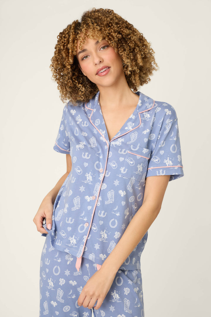 A woman with curly hair models the Whimsy Lone Star Love Cropped PJ Set by P.J. Salvage, featuring blue fabric with white and pink horseshoes and stars, standing against a light background and looking at the camera with a relaxed expression.