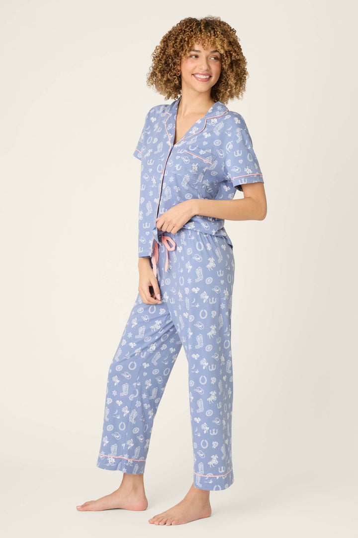 A person with curly hair stands barefoot in P.J. Salvage’s Whimsy Lone Star Love Cropped PJ Set, featuring light blue fabric with white print, pink trim, a short-sleeve button-up top, and long pants. The background is plain and light-colored.