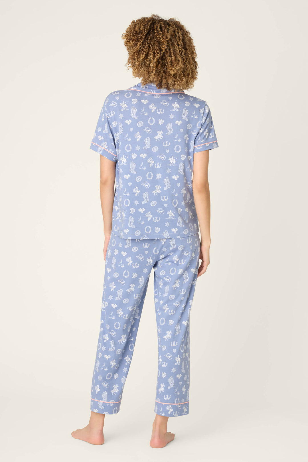 A person with curly hair stands barefoot facing away, wearing the Whimsy Lone Star Love Cropped PJ Set by P.J. Salvage—a matching light blue short-sleeve pajama set with a white nautical print—against a plain, light background.
