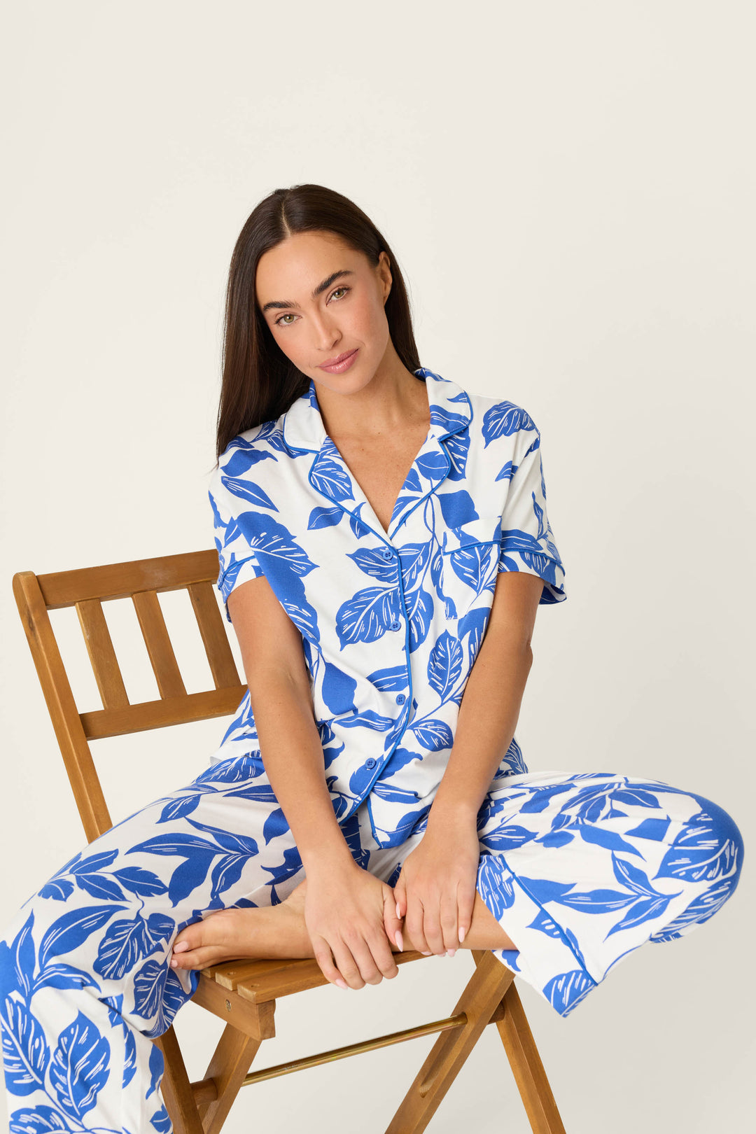 A person with long brown hair sits on a wooden chair, facing the camera in a neutral pose, wearing the P.J. Salvage Whimsy Sapphire Garden Cropped PJ Set featuring a white and blue leaf print, set against a plain background.