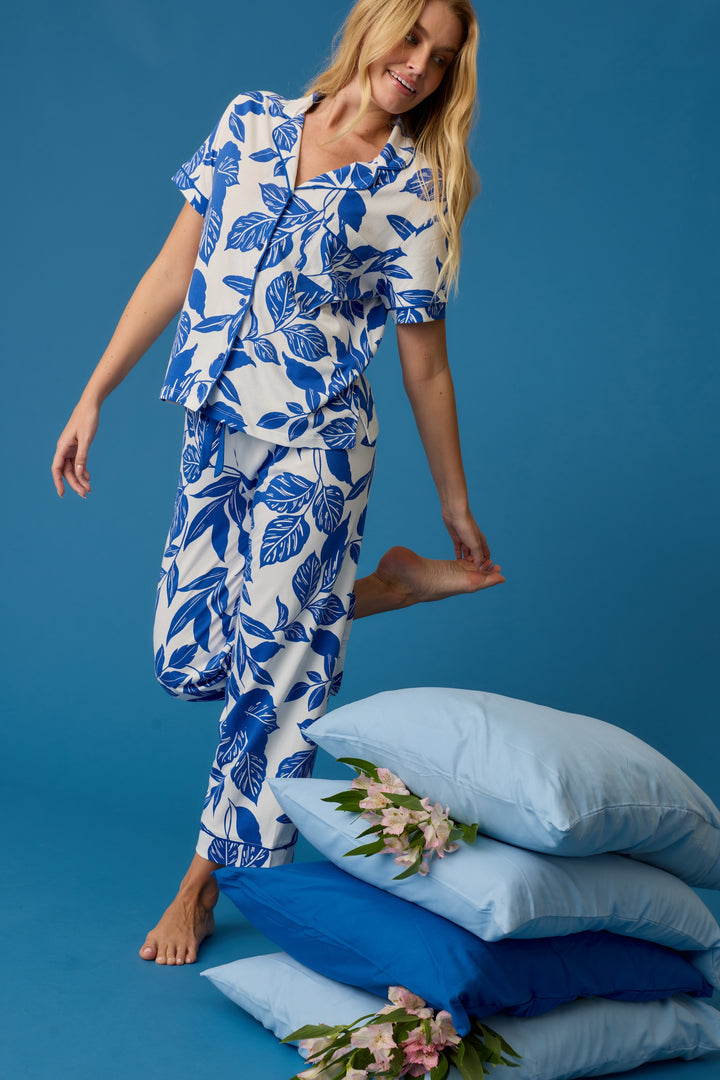 A woman in the P.J. Salvage Whimsy Sapphire Garden Cropped PJ Set poses on one leg beside a stack of blue floral pillows, all set against a solid blue background.