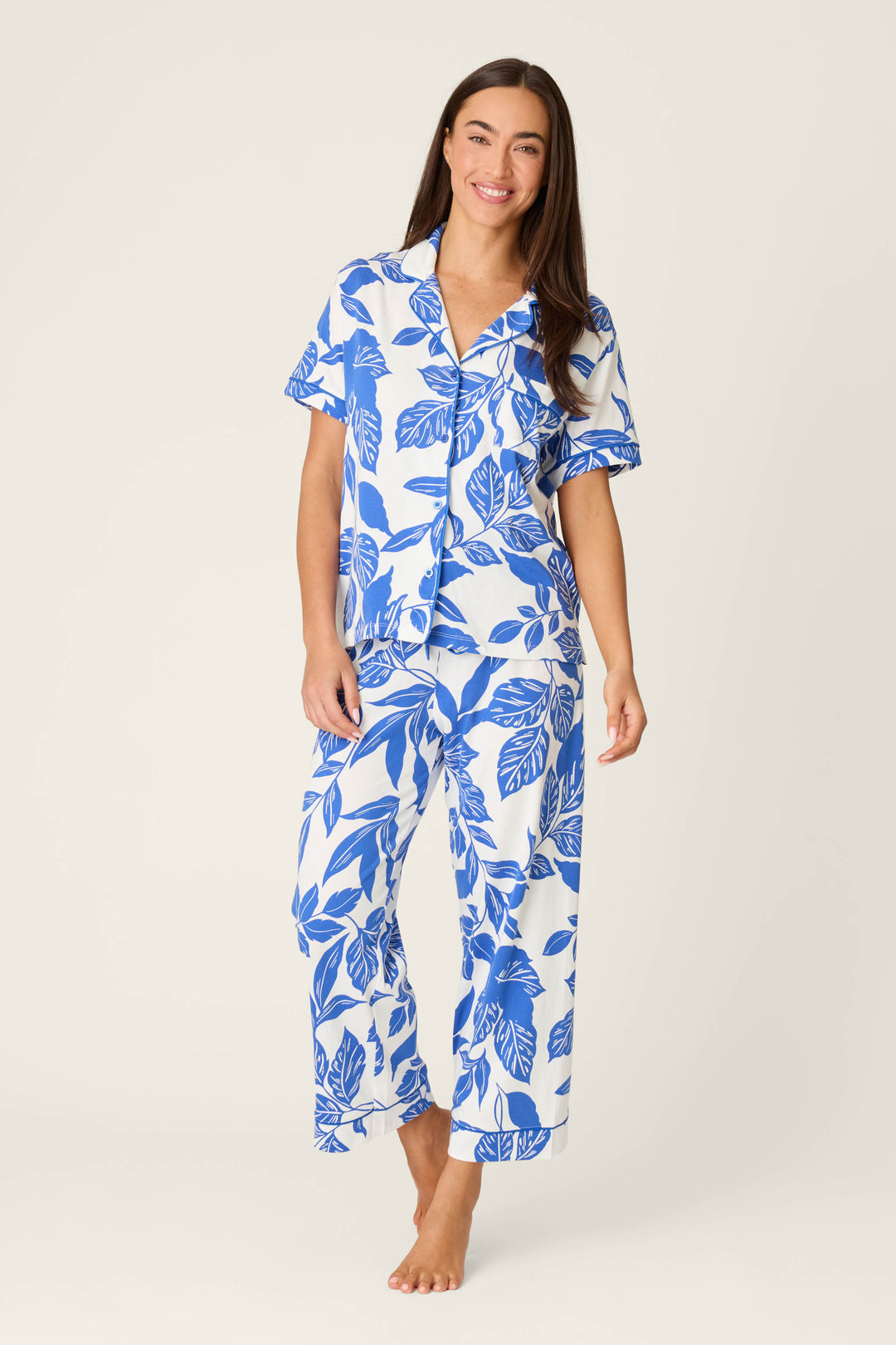 A woman smiles at the camera, barefoot in the Whimsy Sapphire Garden Cropped PJ Set by P.J. Salvage—a matching short-sleeve, button-up shirt and loose-fitting pants featuring a bold blue leaf print on white.