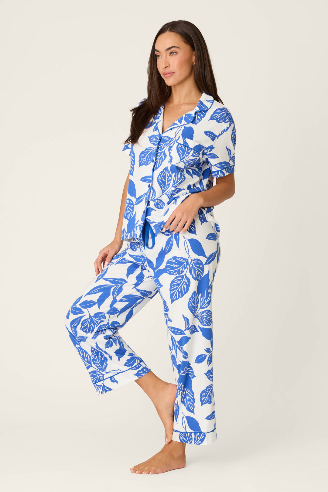 A woman with long brown hair stands barefoot against a plain background, wearing the P.J. Salvage Whimsy Sapphire Garden Cropped PJ Set—a matching short-sleeve pajama set featuring a bold blue leaf print on white fabric. She looks relaxed and neutral.