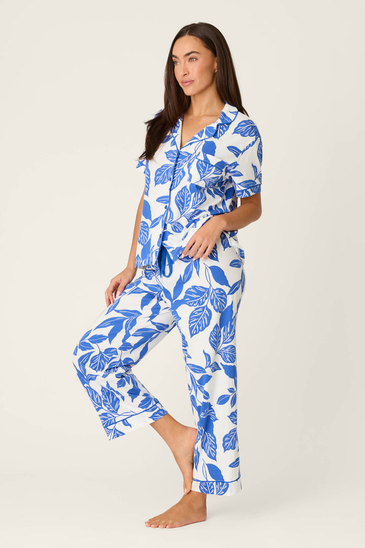 A woman with long brown hair stands barefoot against a plain background, wearing the P.J. Salvage Whimsy Sapphire Garden Cropped PJ Set—a matching short-sleeve pajama set featuring a bold blue leaf print on white fabric. She looks relaxed and neutral.