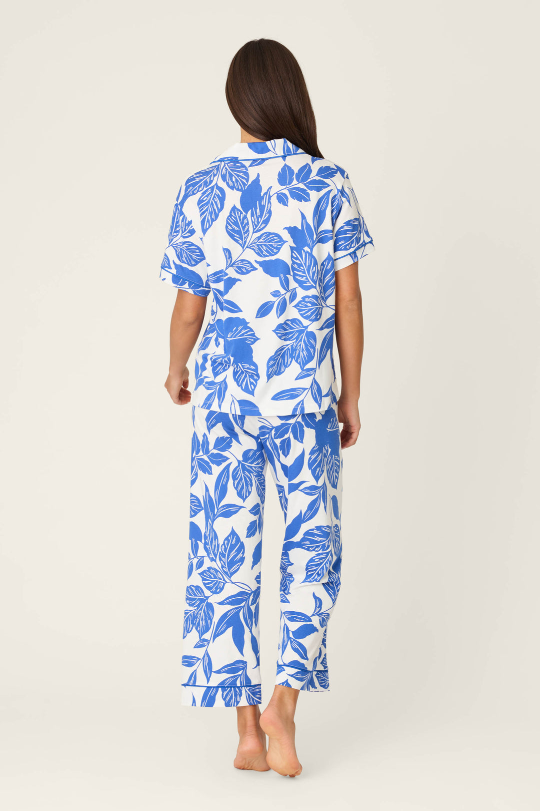 A person with long brown hair stands barefoot, facing away, wearing the P.J. Salvage Whimsy Sapphire Garden Cropped PJ Set—a white pajama set with blue leaf prints and short sleeves. The loose-fitting pants complete the look against a plain off-white background.