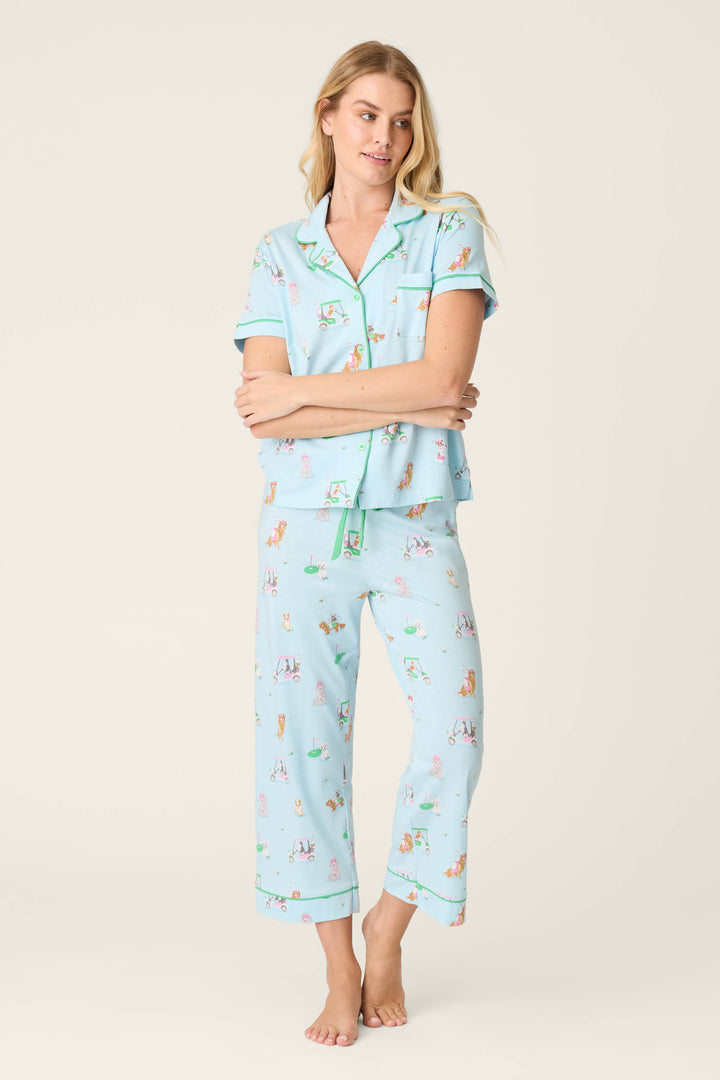 A woman stands barefoot in the P.J. Salvage Whimsy Putter Pup Cropped PJ Set—a light blue pajama featuring a short-sleeve button-up top and long pants with small colorful illustrations—against a plain light background.