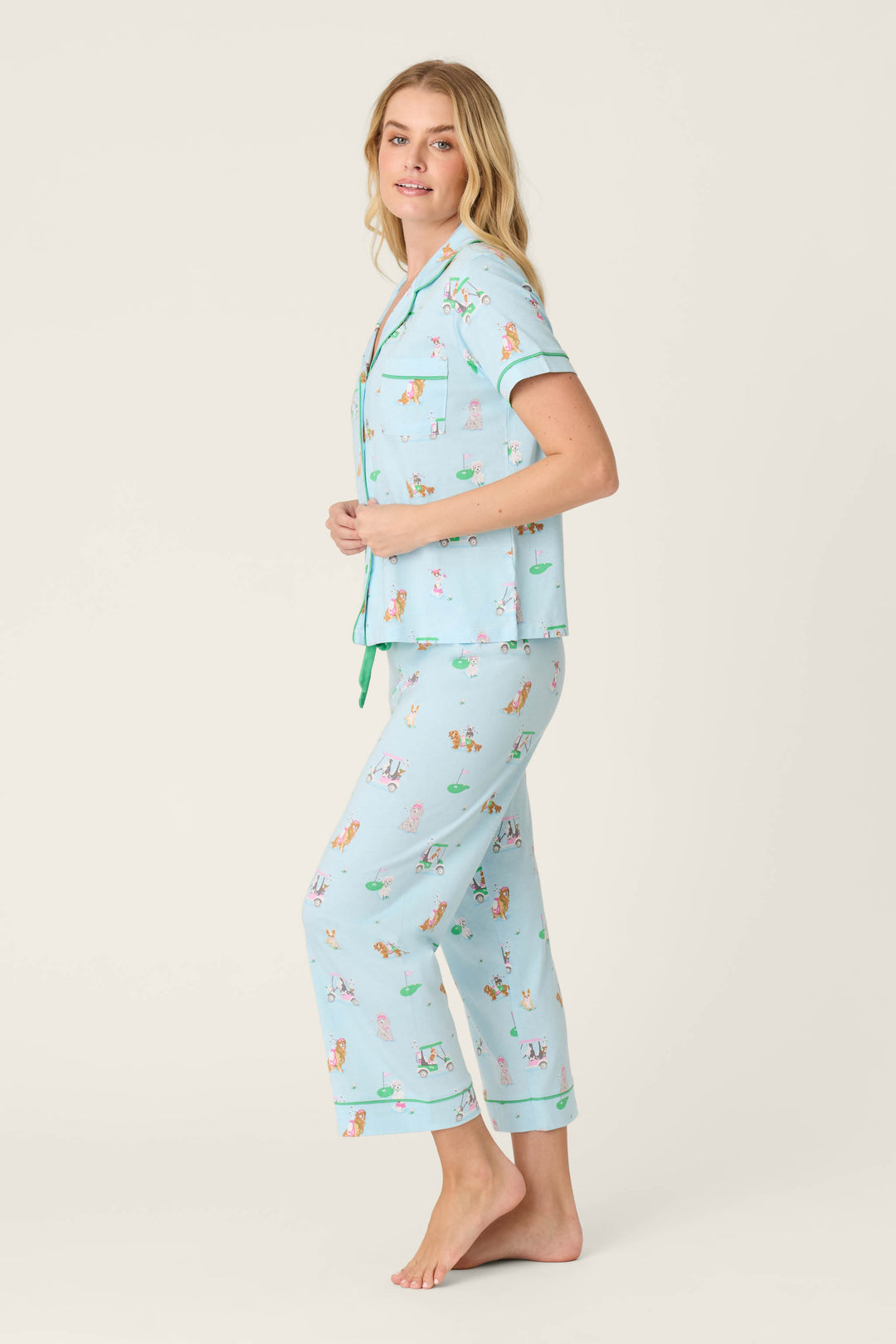 A person stands barefoot against a plain light background, wearing the P.J. Salvage Whimsy Putter Pup Cropped PJ Set—light blue short-sleeve button-up top and long pajama pants with colorful cartoon dog prints.