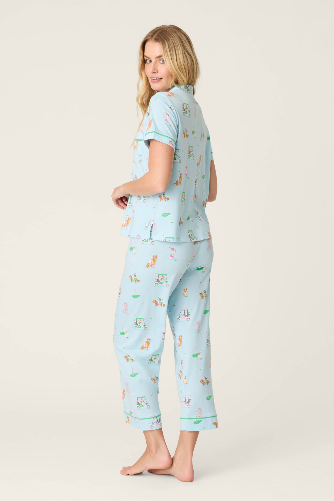 A woman with long blonde hair models the P.J. Salvage Whimsy Putter Pup Cropped PJ Set, featuring a colorful print. She stands barefoot on a plain background, turned slightly to the side and looking over her shoulder.