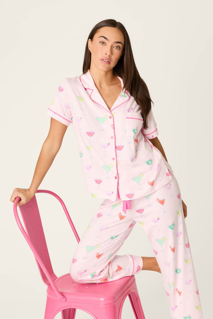 A woman in the P.J. Salvage Whimsy Sip Happens Cropped PJ Set, featuring colorful cocktail glasses, sits on a pink metal chair with one hand on the backrest and her knee bent on the seat.