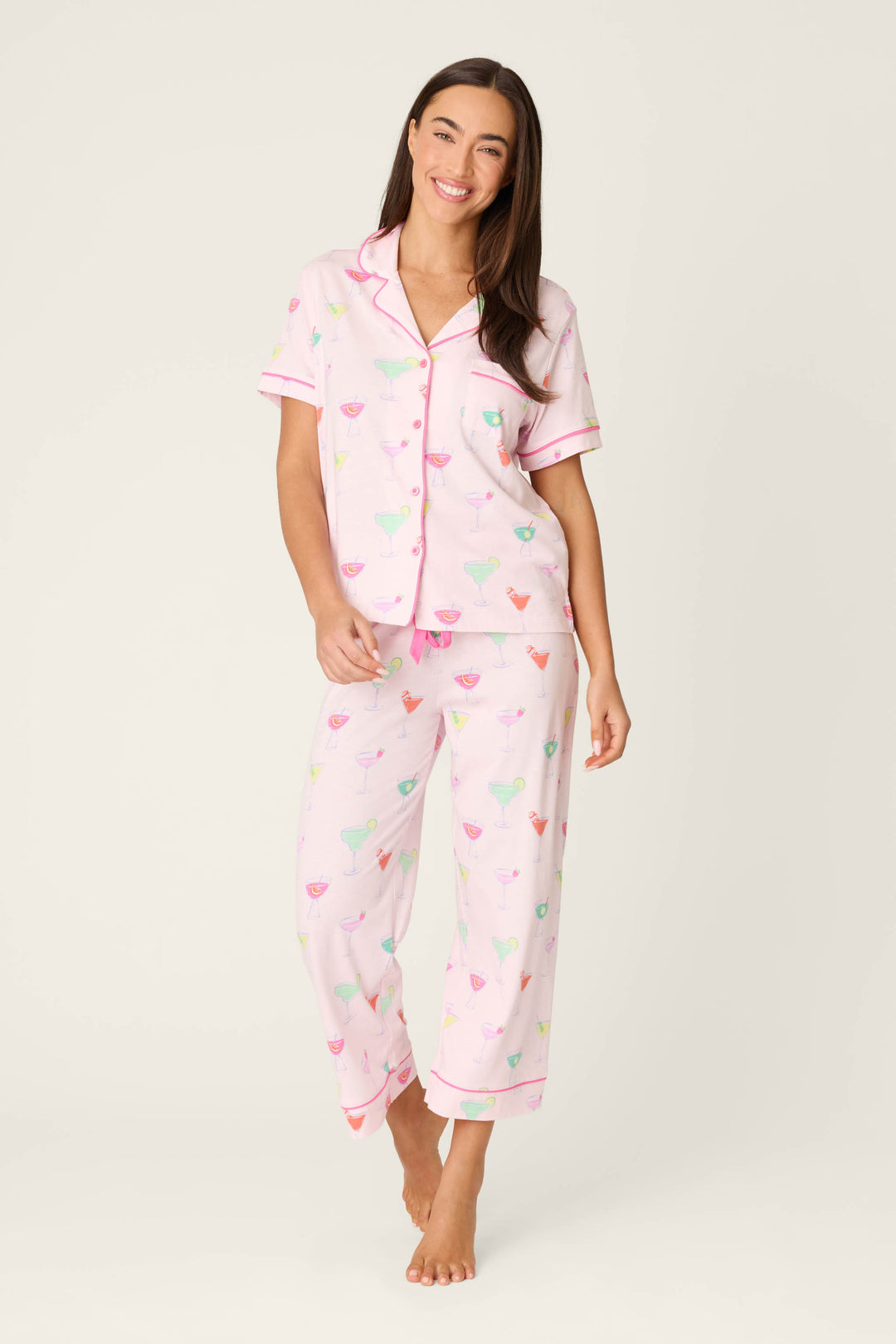 A person stands barefoot against a plain background, wearing the P.J. Salvage Whimsy Sip Happens Cropped PJ Set—light pink pajamas with a colorful cocktail glass print, pink trim, a button-up short-sleeve top, and cropped pants.