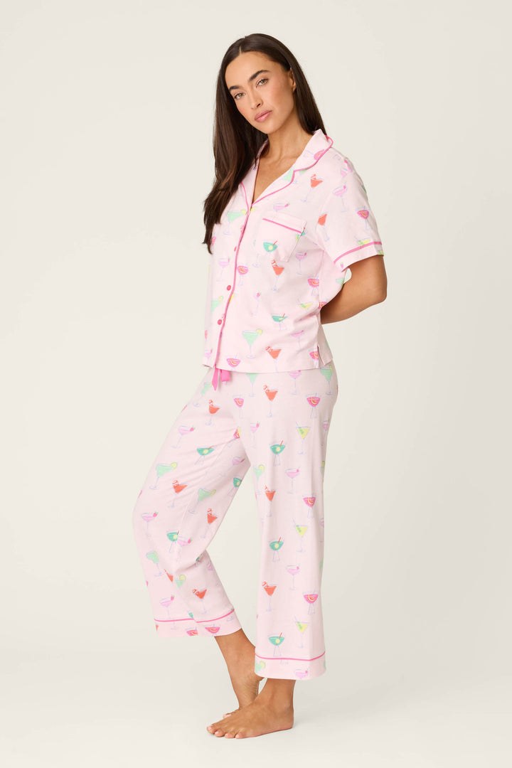 A woman stands barefoot against a plain background, wearing the P.J. Salvage Whimsy Sip Happens Cropped PJ Set—a light pink pajama set with colorful cocktail glass prints, featuring a short-sleeve collared top and cropped pants.