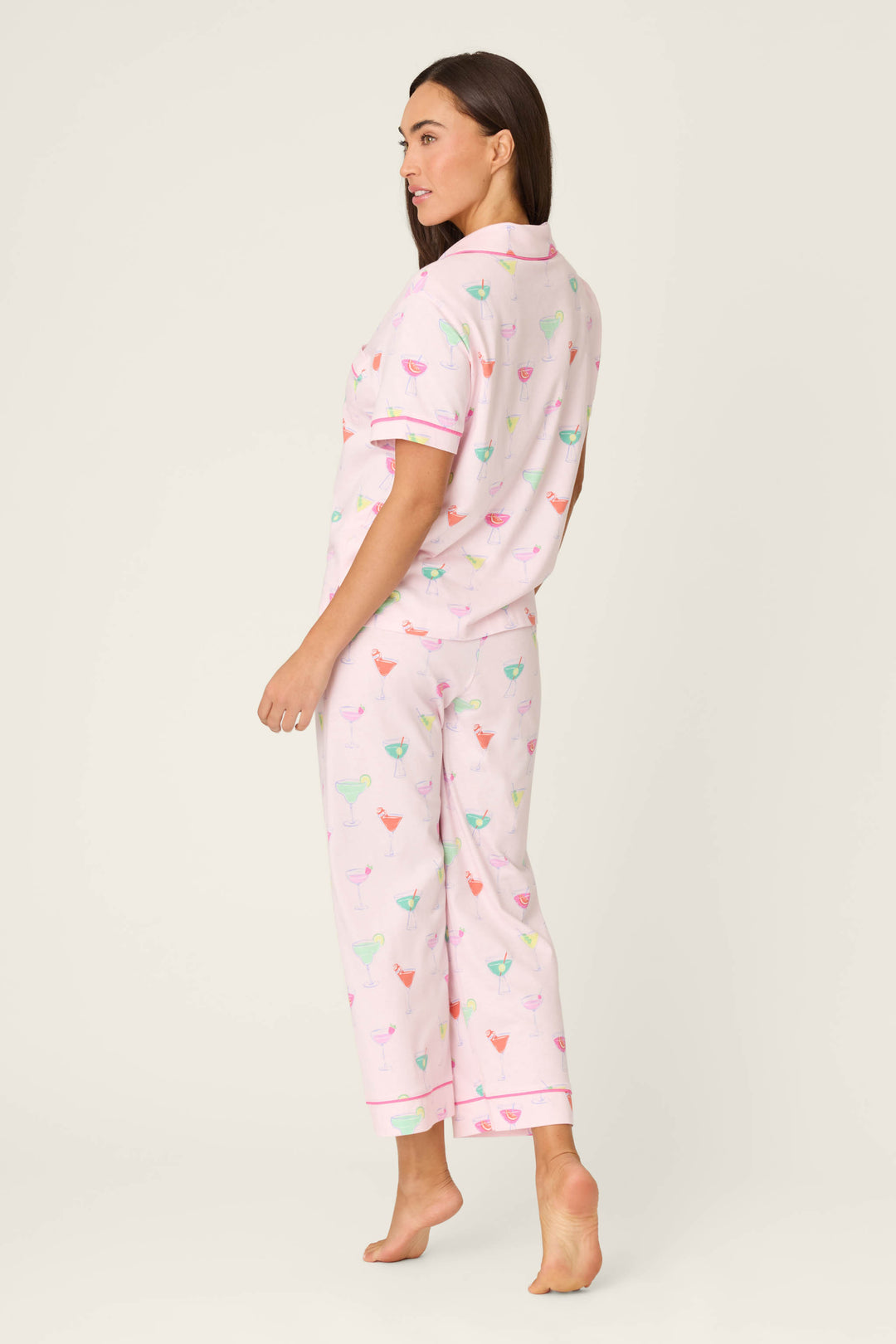 A woman stands barefoot on a plain background, wearing the P.J. Salvage Whimsy Sip Happens Cropped PJ Set featuring a colorful cocktail glass print. She is turned slightly to the side, looking over her shoulder.
