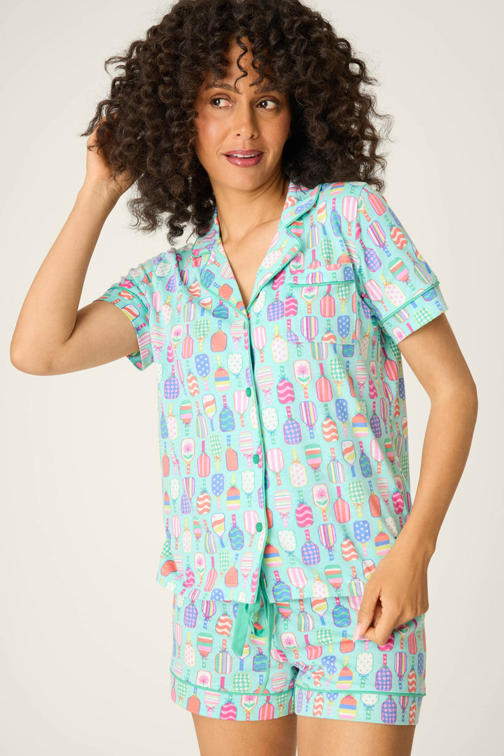 A woman with curly hair wears the P.J. Salvage Whimsy Paddle Party Short PJ Set, featuring a matching short-sleeve button-up top and shorts with a colorful ice cream and popsicle print, standing against a plain background.