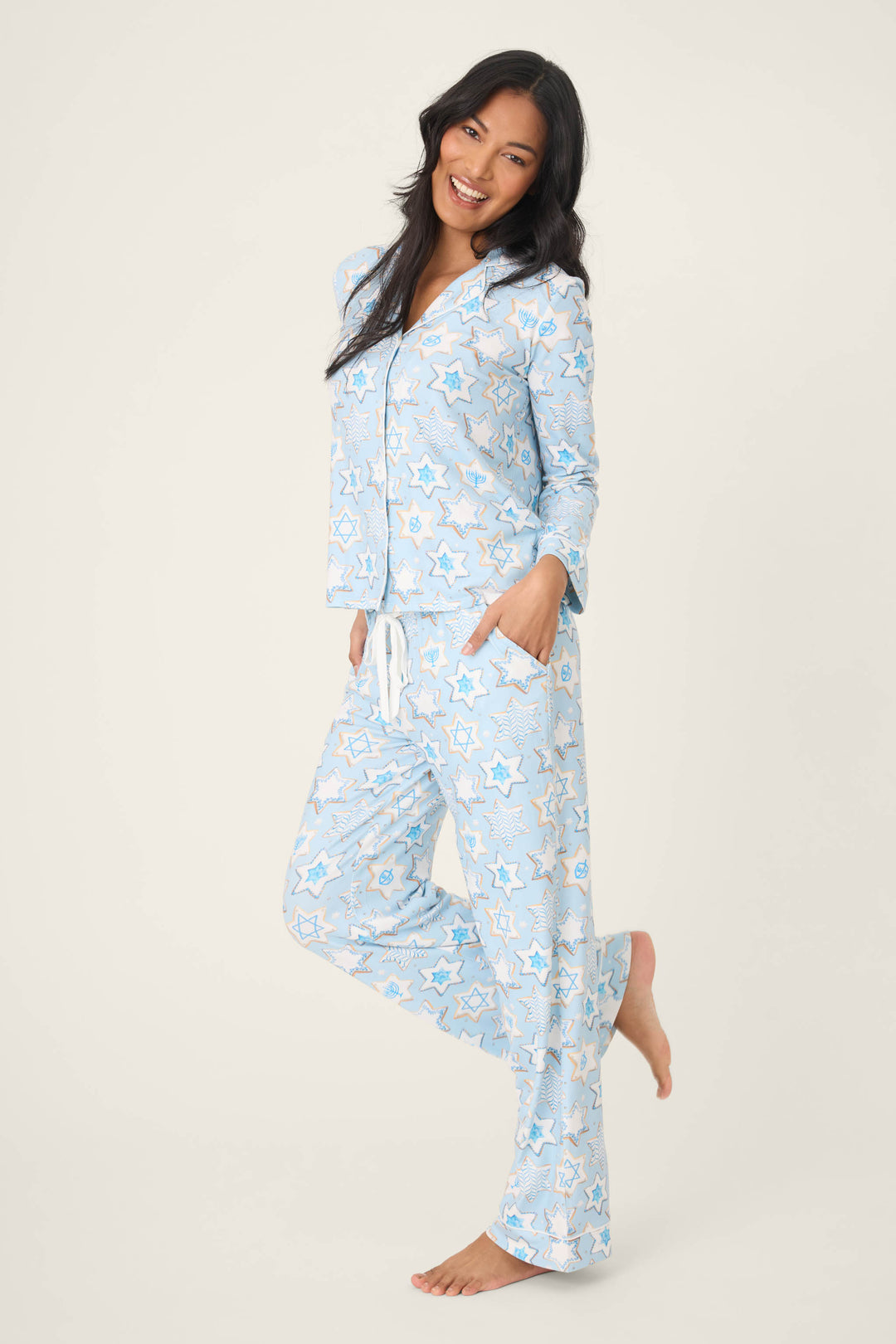 A person stands on one leg, smiling, wearing the HANUKKAH PJ SET YOU HAD ME AT SHALOM by P.J. Salvage—light blue pajamas with a star pattern, long sleeves, buttons, pants with pockets and drawstring—against a plain light background.