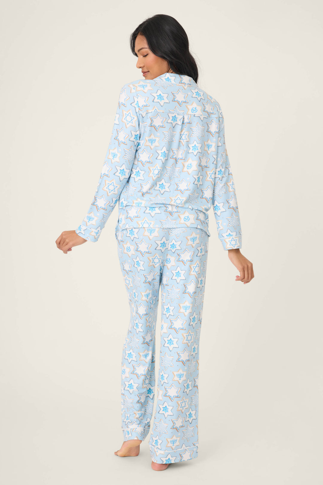 A woman with long dark hair stands barefoot, facing away, wearing the P.J. Salvage HANUKKAH PJ SET YOU HAD ME AT SHALOM—light blue pajamas with a white and yellow star pattern—her right hand slightly extended behind her.