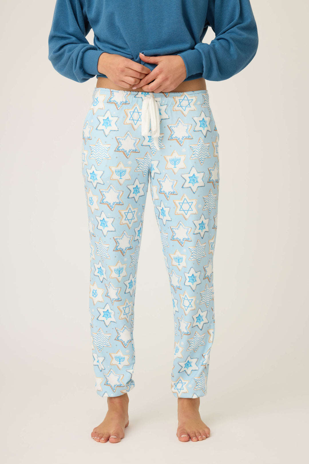 A person wears P.J. Salvages HANUKKAH BANDED PANT YOU HAD ME AT SHALOM—light blue pajama pants featuring Star of David and menorah patterns—standing barefoot against a plain background, with their upper body partially visible.