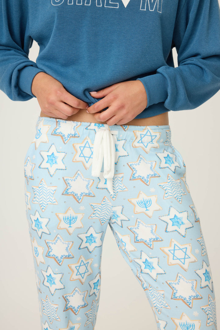 A person wears P.J. Salvages HANUKKAH BANDED PANT YOU HAD ME AT SHALOM—light blue pajama pants with Star of David and menorah patterns—paired with a blue sweatshirt, adjusting the waistband.
