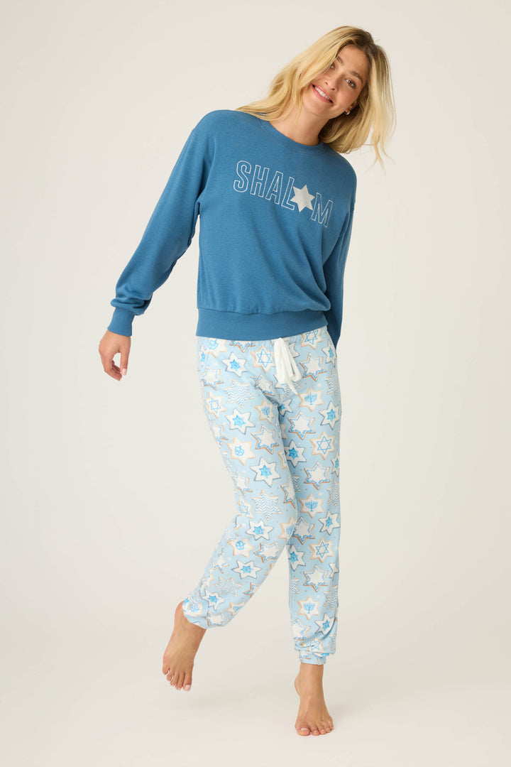 A woman stands barefoot and smiling against a plain light background, wearing P.J. Salvage’s HANUKKAH BANDED PANT YOU HAD ME AT SHALOM pajama pants with blue and yellow stars, and a blue “SHALOM” sweatshirt with a star.