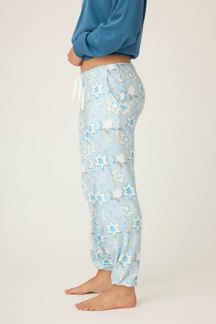 A person stands barefoot and sideways on a plain light background, wearing P.J. Salvages HANUKKAH BANDED PANT YOU HAD ME AT SHALOM featuring a Star of David pattern and light blue color with a blue long-sleeve shirt.