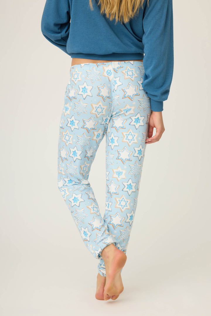 A person with long blonde hair stands barefoot, facing away, wearing P.J. Salvage HANUKKAH BANDED PANT YOU HAD ME AT SHALOM pajama pants in light blue with Stars of David and menorah patterns, paired with a blue top.