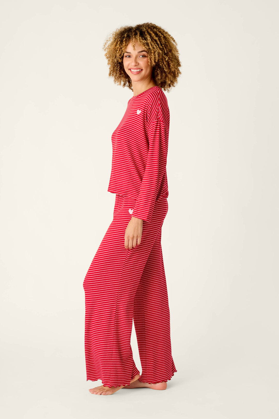A woman smiles wearing an Icon Stripes Red Long Sleeve PJ Set, a red pajama set with thin white horizontal stripes, small white heart appliqués, a long-sleeved shirt, and matching wide-leg pants.