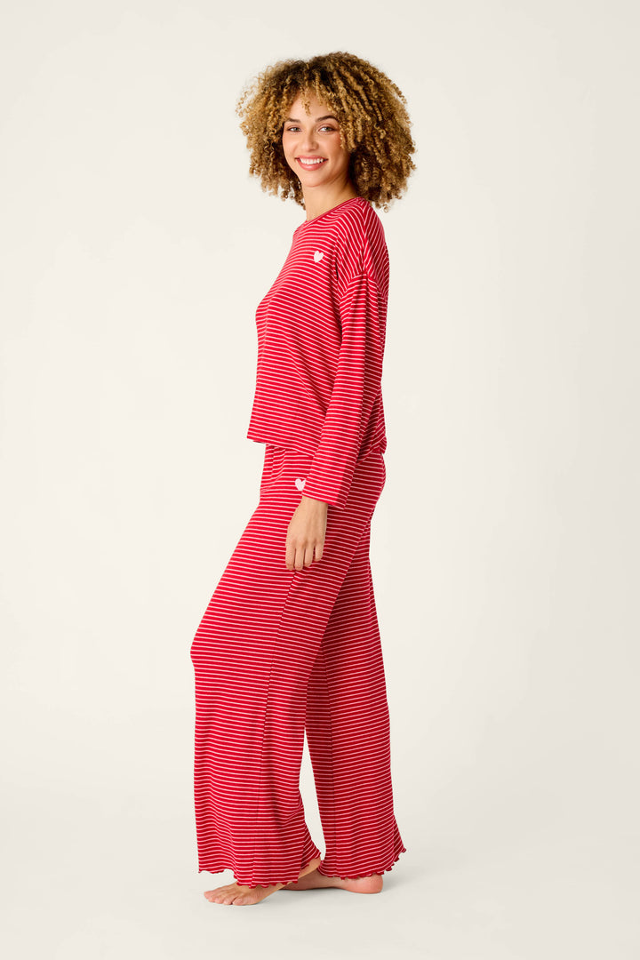 A woman smiles wearing an Icon Stripes Red Long Sleeve PJ Set, a red pajama set with thin white horizontal stripes, small white heart appliqués, a long-sleeved shirt, and matching wide-leg pants.
