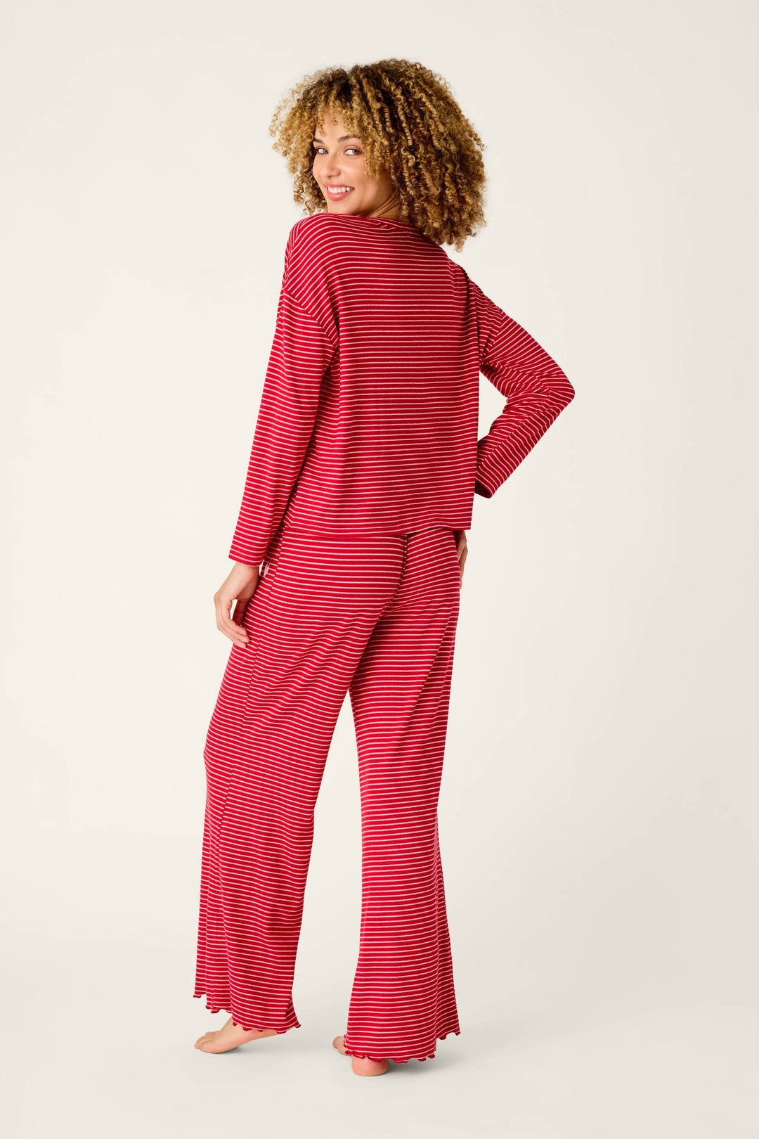 A woman with curly hair wearing an Icon Stripes Red Long Sleeve PJ Set, featuring a red and white fine horizontal stripe long-sleeved top and matching pants.