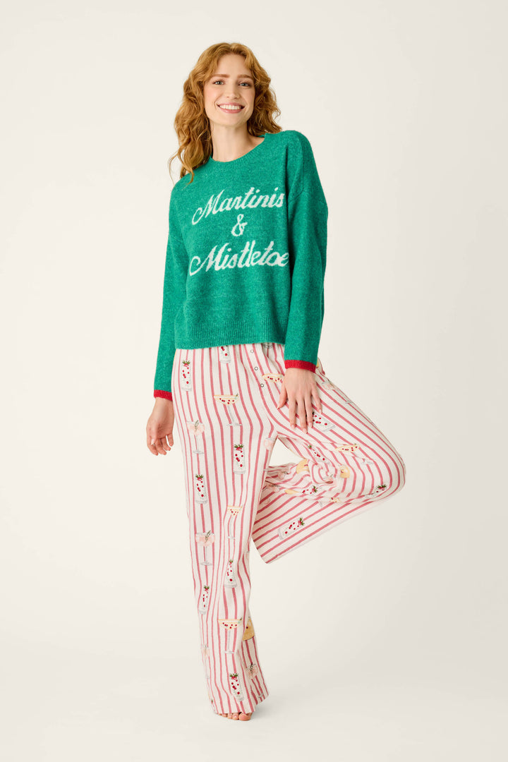 A smiling woman wears a festive P.J. Salvage Martinis & Mistletoe Snuggle Sweater and red-and-white striped pajama pants featuring a martini glass print.