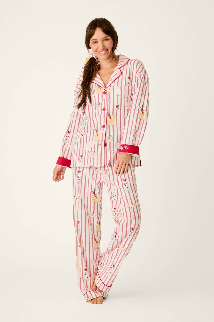 A woman wears P.J. Salvage Martinis & Mistletoe Cocktail Print Flannel 3-Piece PJ Set pajamas, consisting of a button-down top and matching trousers in a red-and-white vertical stripe pattern with cocktail glass and mistletoe illustrations.