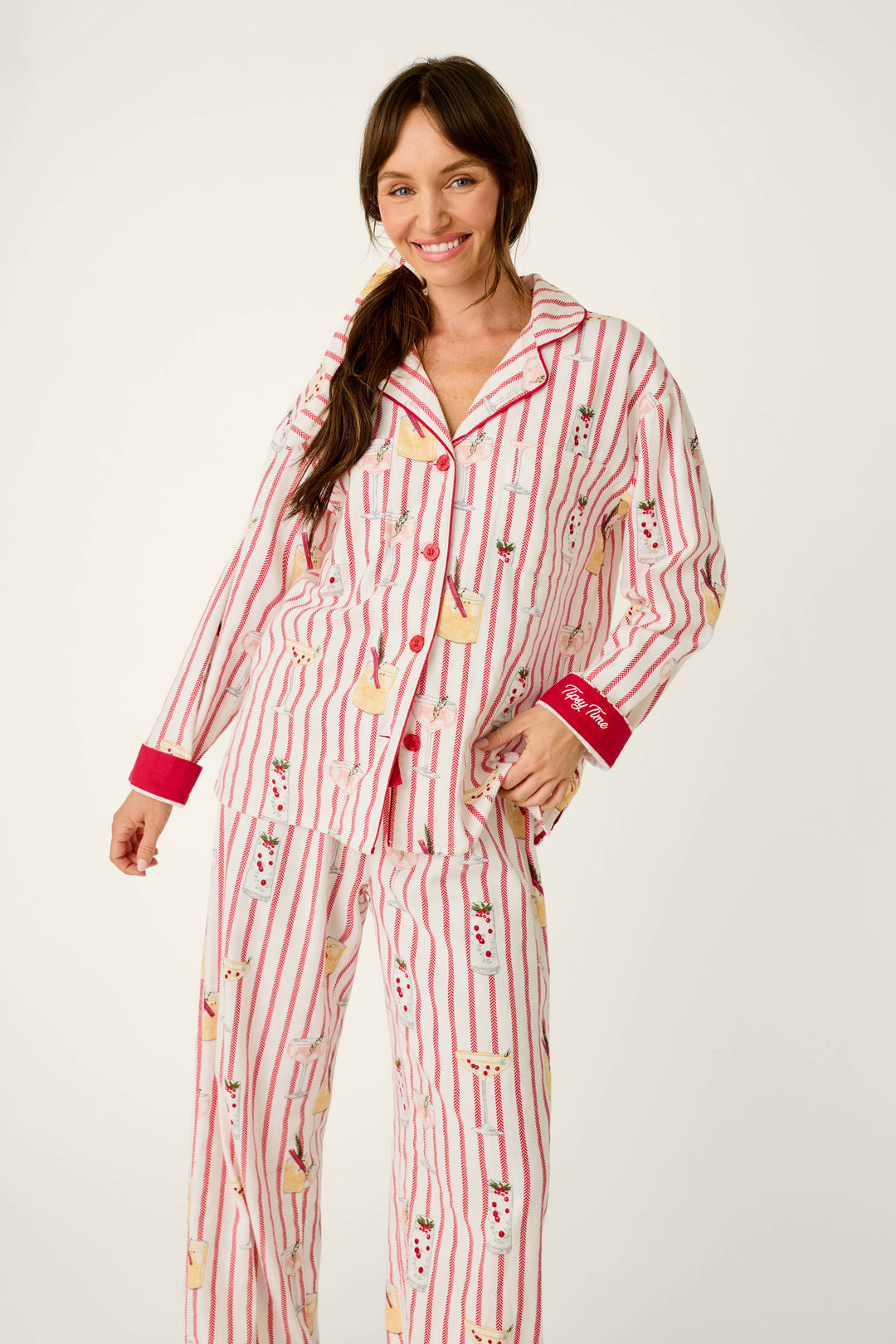 A woman wears a P.J. Salvage Martinis & Mistletoe Cocktail Print Flannel 3-Piece PJ Set, featuring a cozy white and red striped pattern with cocktail illustrations, red trim, and Tipsy Time embroidered on the sleeve cuff.