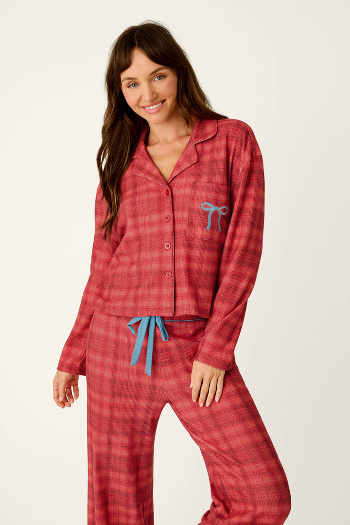 A smiling woman wears P.J. Salvage Mad for Plaid Long Sleeve Notch Collar Top red plaid waffle knit pajamas, featuring a button-down top with a blue chest-pocket bow and matching pants with a blue drawstring.