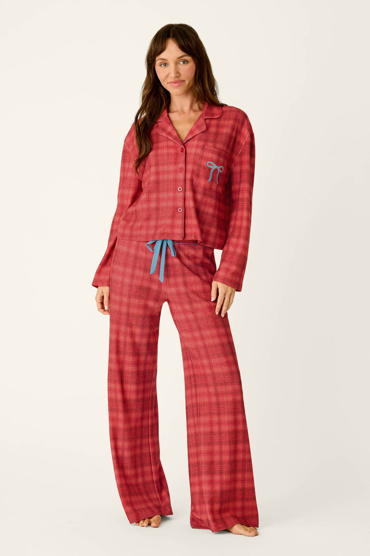 A woman wears the P.J. Salvage Mad for Plaid Long Sleeve Notch Collar Top and matching wide-leg pants, featuring a red plaid pattern, blue embroidered bow detail on the pocket, and a blue drawstring waist.
