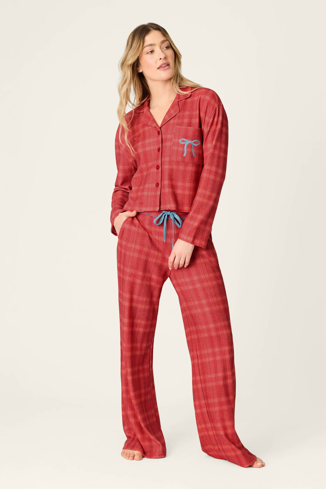 A woman wearing a P.J. Salvage Mad for Plaid Long Sleeve & Pants PJ Set in red plaid with a blue bow detail on the pocket and a blue drawstring waist.