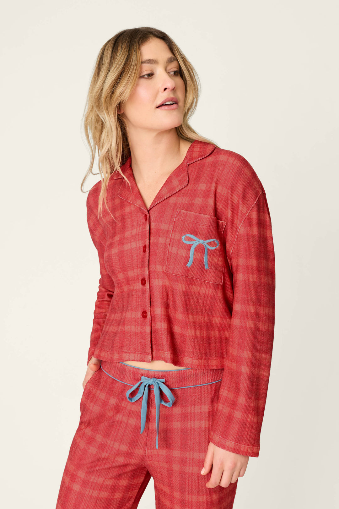 A woman wearing a P.J. Salvage Mad for Plaid Long Sleeve & Pants PJ Set in red plaid with light blue bow detailing, long sleeves, and a drawstring waist.