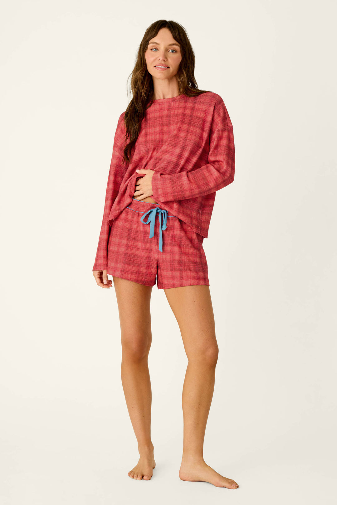 A woman wearing a P.J. Salvage Mad for Plaid Long Sleeve & Shorts PJ Set, featuring a cozy red plaid long-sleeve top and matching shorts with a blue drawstring waist.
