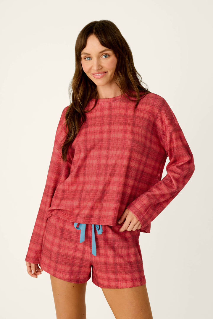 A woman wearing a P.J. Salvage Mad for Plaid Long Sleeve & Shorts PJ Set, consisting of a long-sleeve top and shorts in a red plaid print with a blue drawstring waist.