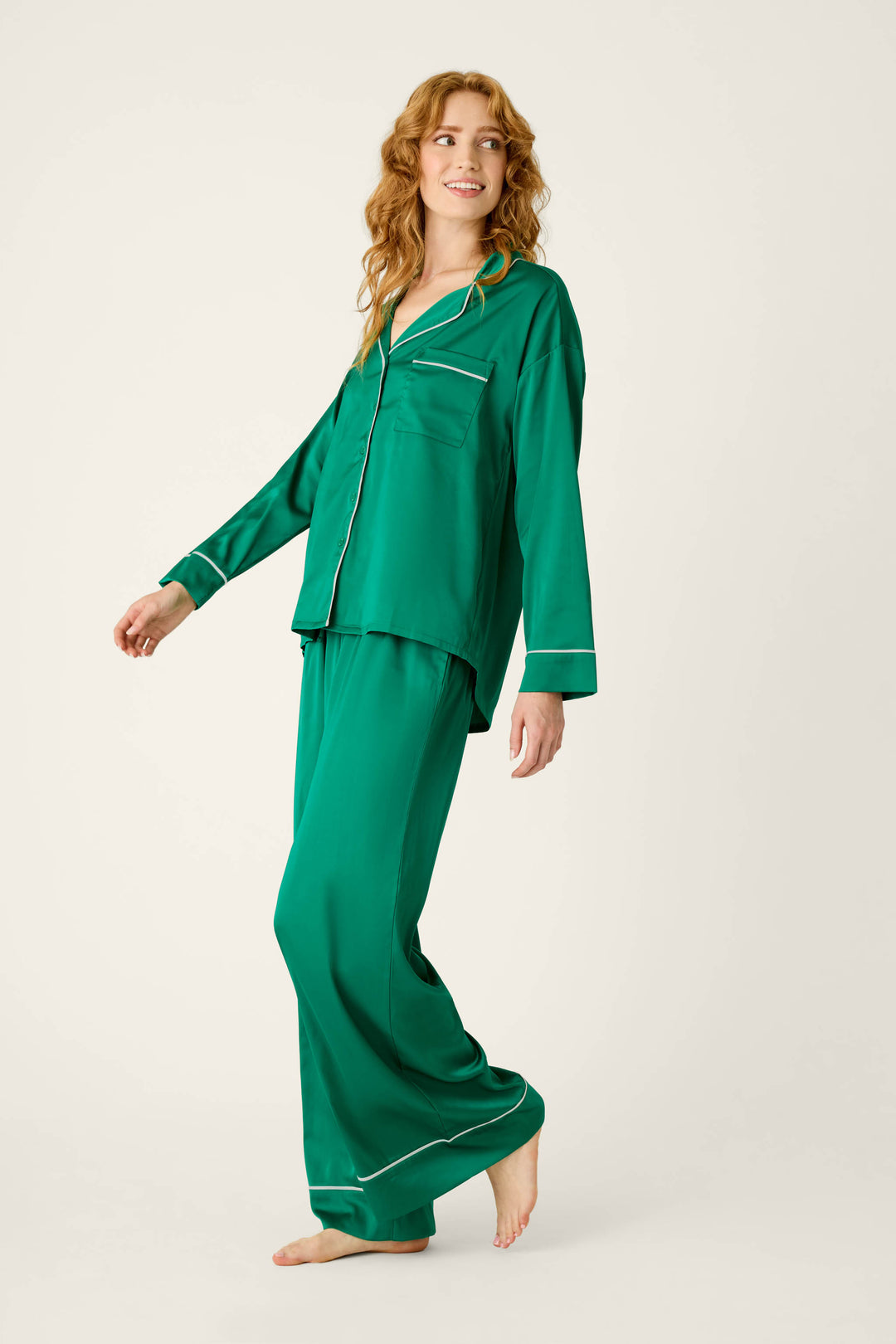 A woman wearing a P.J. Salvage Moonlight Muse Green Satin Long Sleeve PJ Set, featuring a luxe green satin long-sleeved button-down top and matching wide-leg pants with white piping.