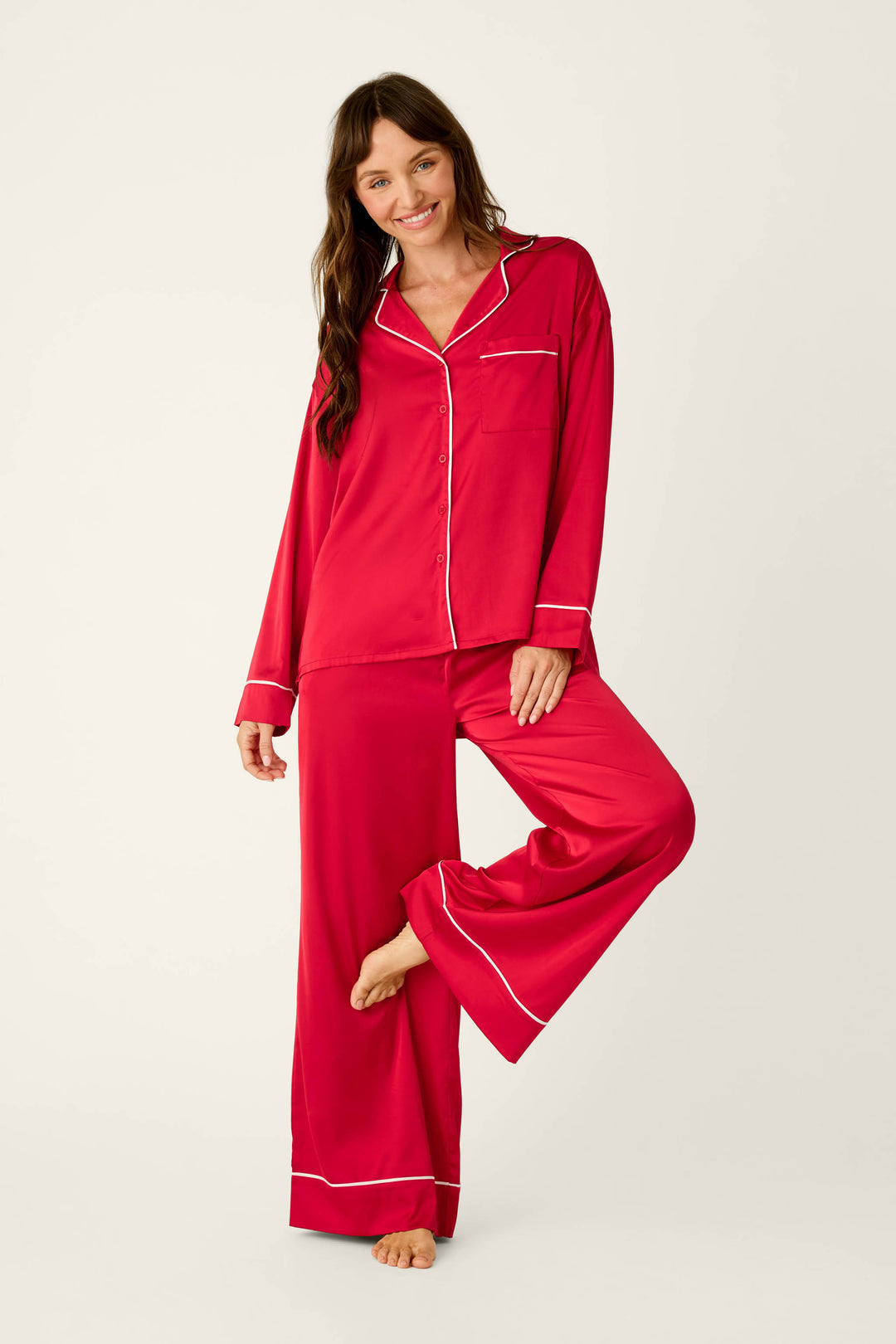 A woman wears a P.J. Salvage Moonlight Muse Red Satin Long Sleeve PJ Set, featuring a luxe red button-up top and matching wide-leg pants with white piping trim.