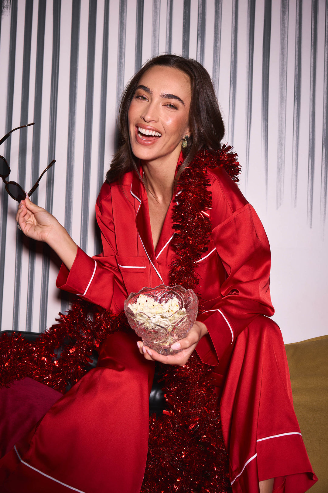 A smiling woman wears a Moonlight Muse Red Satin Long Sleeve PJ Set with white piping, holding a bowl of popcorn and a pair of sunglasses, with red tinsel draped around her.