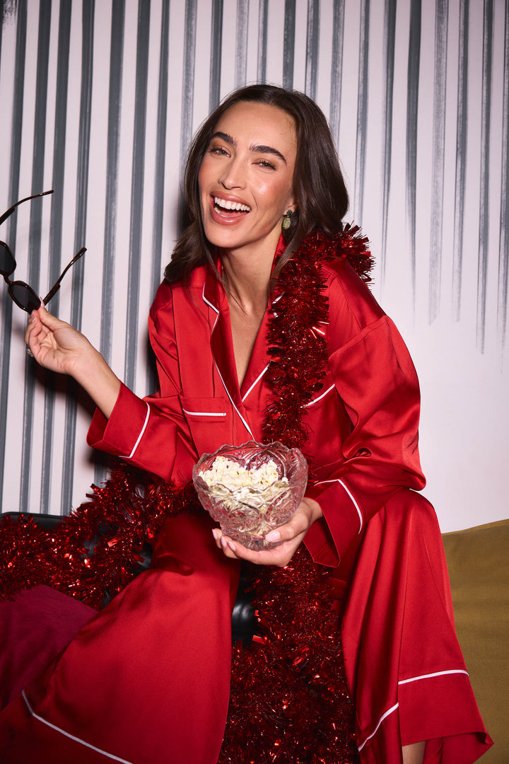 A smiling woman wears a Moonlight Muse Red Satin Long Sleeve PJ Set with white piping, holding a bowl of popcorn and a pair of sunglasses, with red tinsel draped around her.