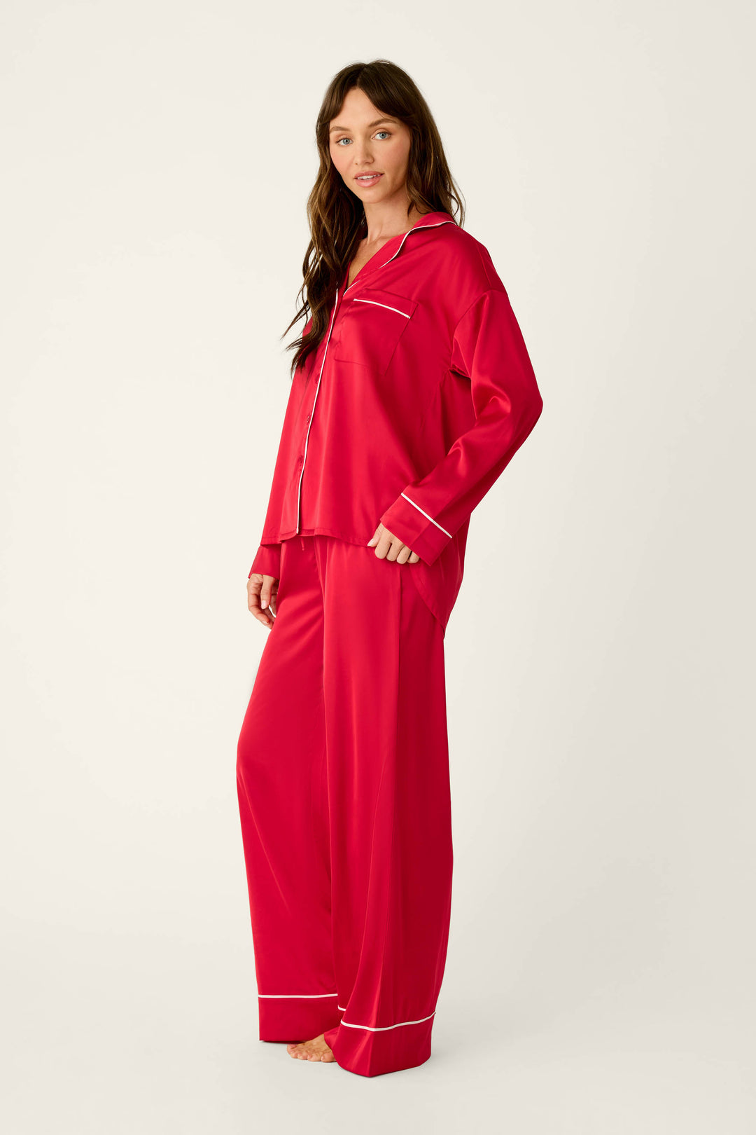 A person wearing a P.J. Salvage Moonlight Muse Red Satin Long Sleeve PJ Set, consisting of a long-sleeved button-down top and matching wide-leg trousers with white piping trim.