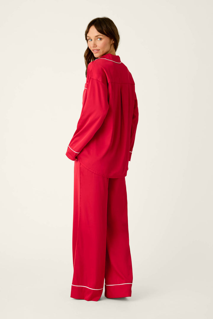 A woman wearing a P.J. Salvage Moonlight Muse Red Satin Long Sleeve PJ Set featuring a long-sleeved button-down top and matching wide-leg pants with white piping trim.