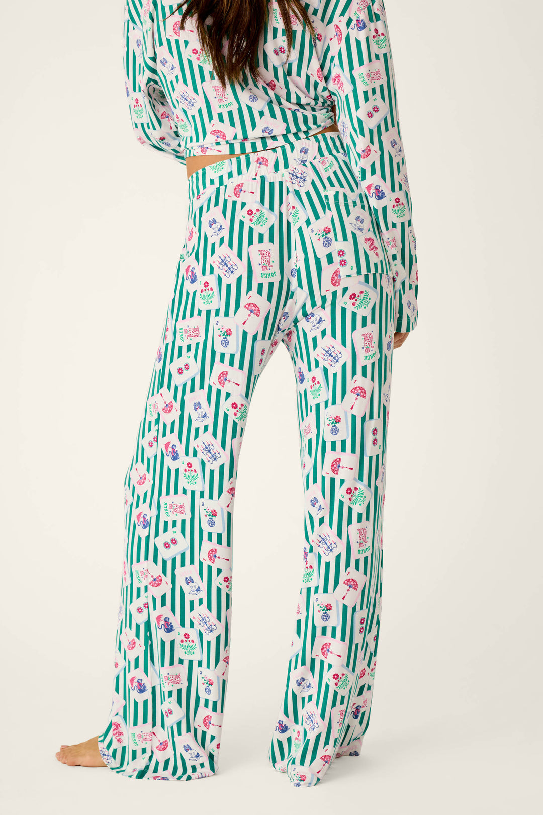 A person wearing P.J. Salvage Mahjong Queen Dreams Pajama Pants and a long-sleeved top featuring a green-and-white vertical striped pattern decorated with playing card motifs.