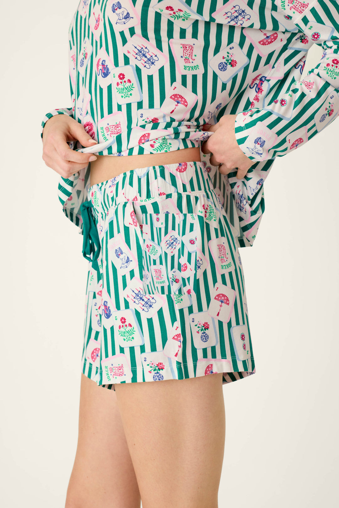 Person wearing P.J. Salvage Mahjong Queen Dreams Pajama Shorts, a matching long-sleeved top in modal jersey, both featuring a pattern of green vertical stripes and colorful mahjong tiles.