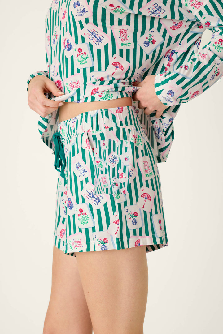 Person wearing P.J. Salvage Mahjong Queen Dreams Pajama Shorts, a matching long-sleeved top in modal jersey, both featuring a pattern of green vertical stripes and colorful mahjong tiles.