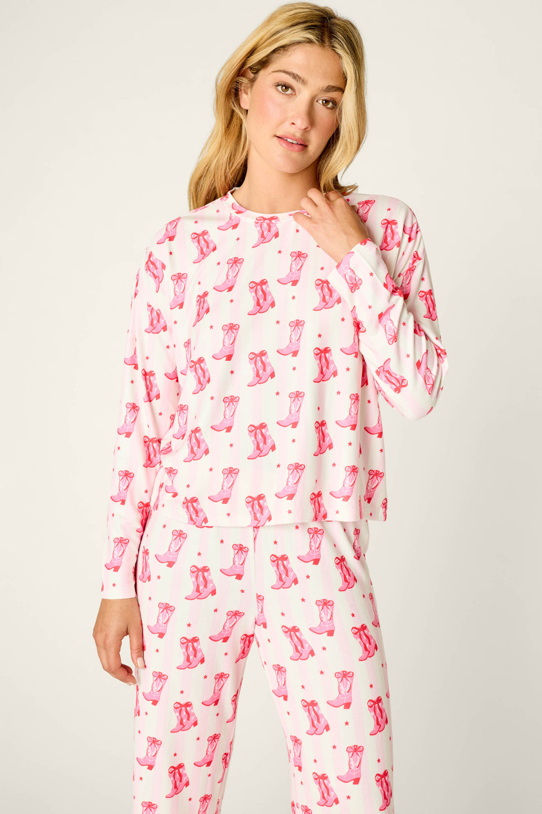 A woman wears the P.J. Salvage Midnight Rodeo Cowboy Boots & Stripe Print Long Sleeve PJ Set, featuring a floral top and blue pants against a pink-and-white striped background with pink cowboy boots and small stars.