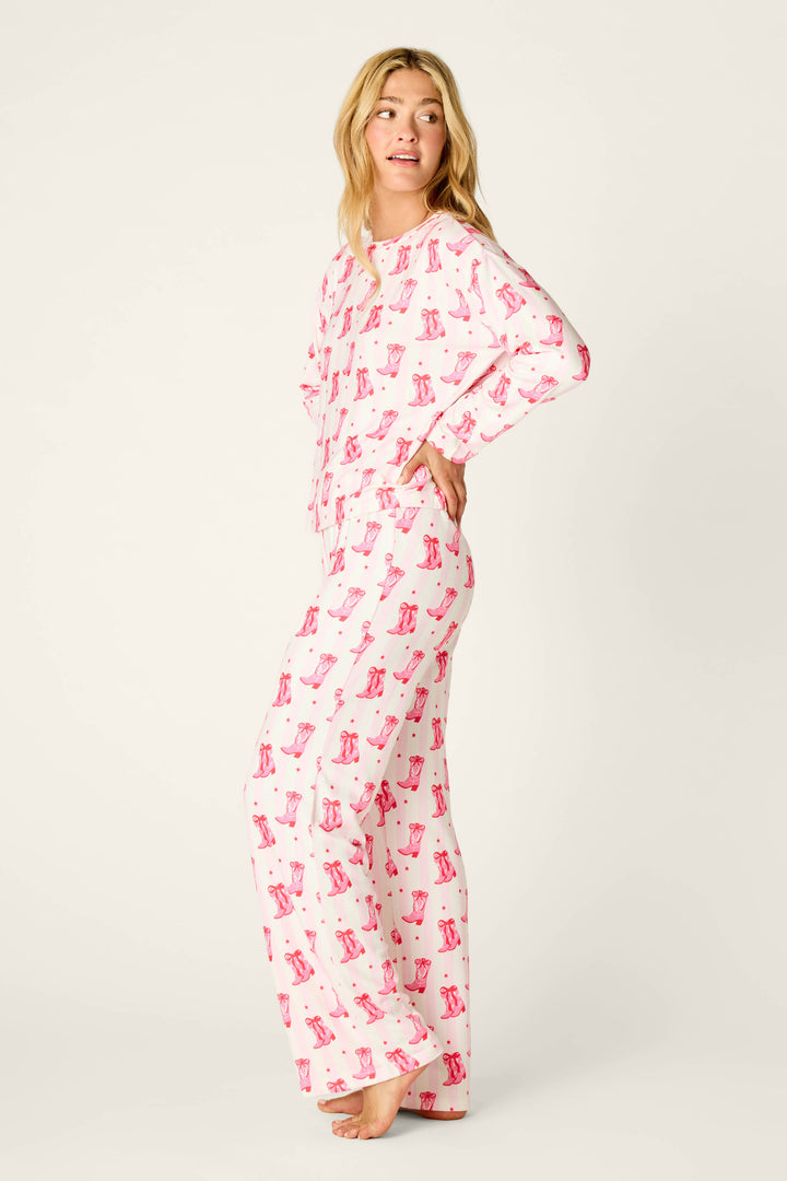 A woman wears the P.J. Salvage Midnight Rodeo Cowboy Boots & Stripe Print Long Sleeve PJ Set, a double-brushed pointelle pajama set with a long-sleeved top and wide-leg trousers patterned with small pink cowboy boots and red hearts.