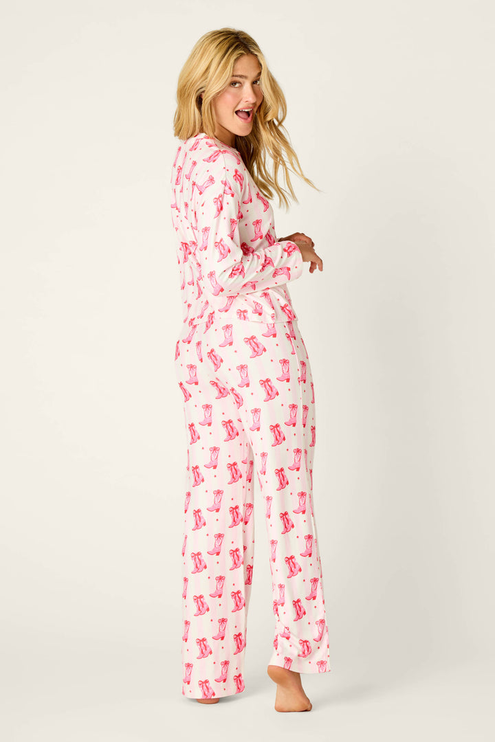 A woman wears the P.J. Salvage Midnight Rodeo Cowboy Boots & Stripe Print Long Sleeve PJ Set, featuring a pink cowboy boot and polka dot print on a white striped background.