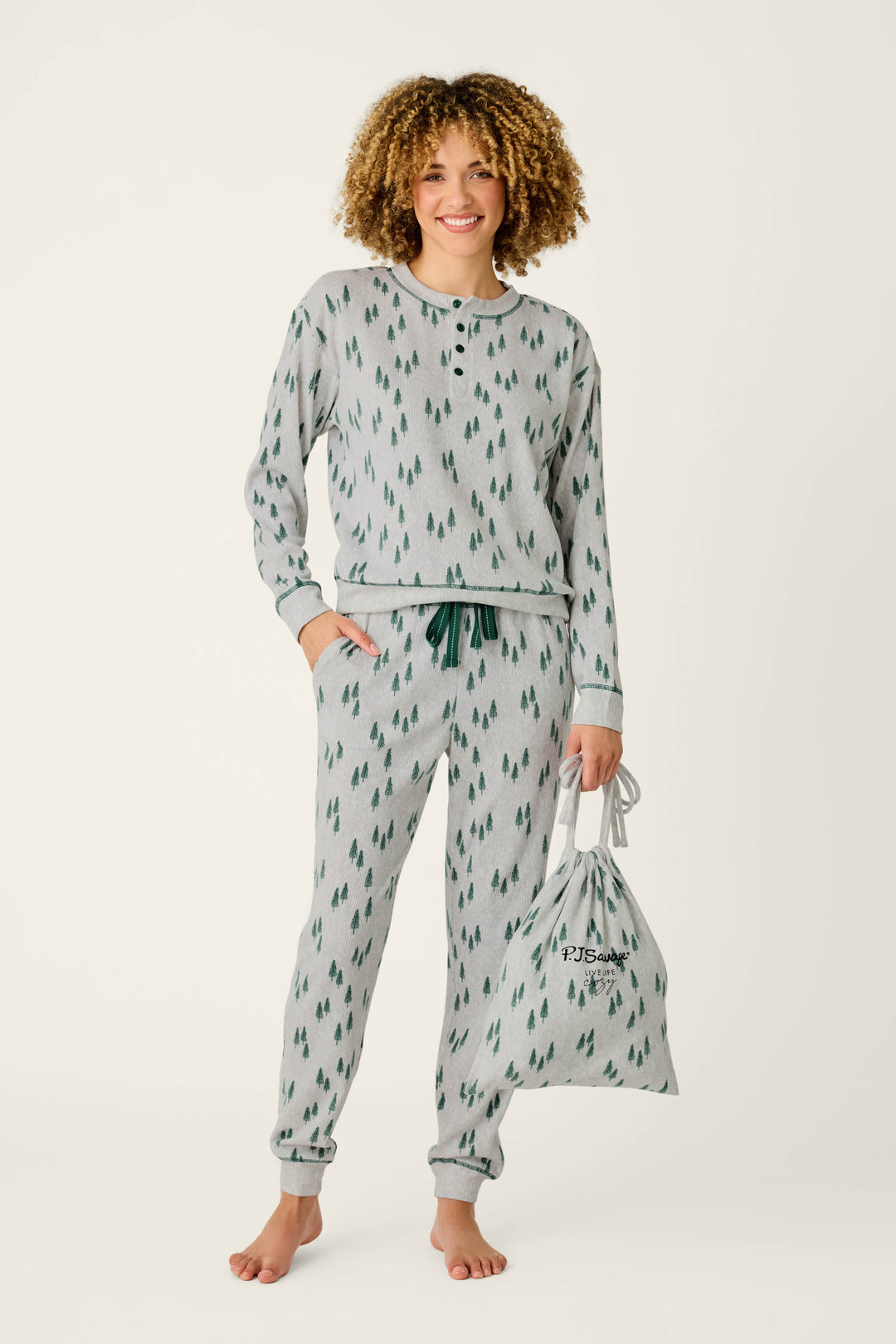 A smiling person with curly hair stands barefoot against a plain background, wearing the P.J. Salvage Trees Print 3-Piece PJ Gift Set—gray pajamas featuring a green tree pattern—while holding the matching drawstring bag.
