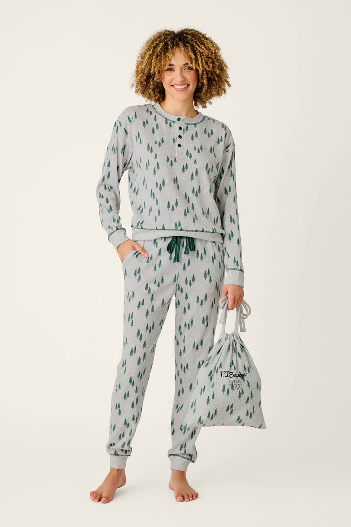 A smiling person with curly hair stands barefoot against a plain background, wearing the P.J. Salvage Trees Print 3-Piece PJ Gift Set—gray pajamas featuring a green tree pattern—while holding the matching drawstring bag.